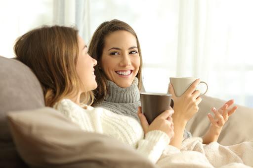 Two women wearing sweaters sitting on a couch with cups of coffee.